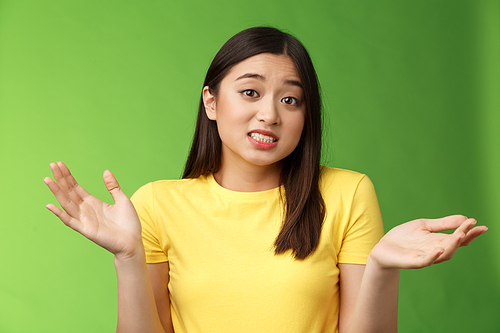 Confused cute apologizing asian girl shrugging hands spread sideways, clench teeth feel sorry, perplexed answer, cannot say, unsure and uncertain, clueless what happened, green background.