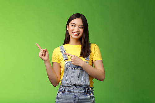 Lovely pretty tender asian woman showing way, tilt head flirty smiling, helpful pointing left, introduce plan, gladly present promo recommend cool new product, stand green background happy.