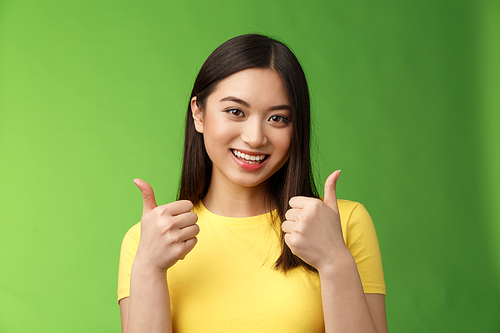 Supportive cute asian girlfriend rooting for lover show thumbs-up approval sign, joyfully smiling, like good choice, accept excellent effort, encourage friend, stand green background. Copy space