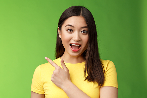 Enthusiastic impressed cute asian brunette describing new haircare product features amused and amazed, lively talking poining left astonished, have interesting discussion, stand green background.