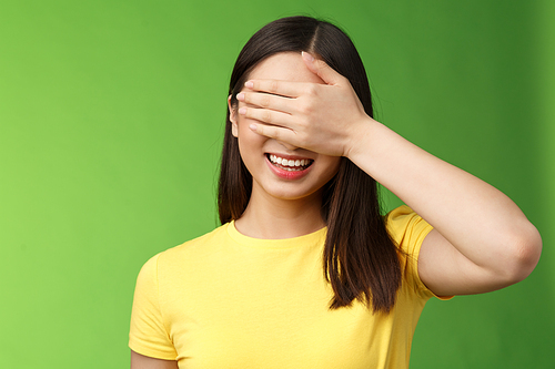 Close-up cheerful amused tender asian girlfriend close eyes waiting friend give surprise gift, smiling broadly hold palm face, anticipating interesting present, stand green background intrigued.