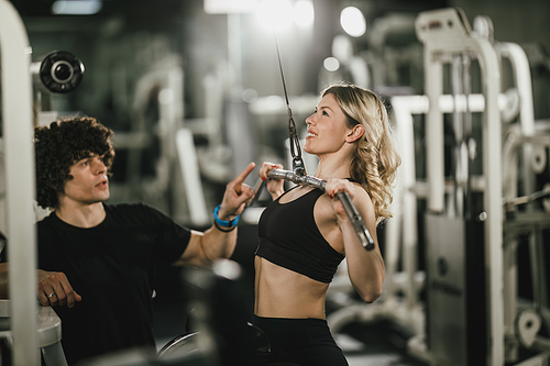 A young muscular woman is doing training at machine supporting by coach in the gym.