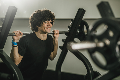 Shot of a muscular guy working out at the hard training in a gym. He is doing squat exercises with barbell.