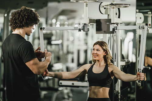 A young muscular woman is doing training at machine supporting by coach in the gym.