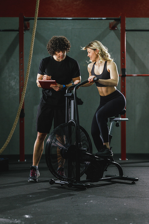 A young muscular woman is doing hard cal bike crossfit training in the gym supporting by coach.