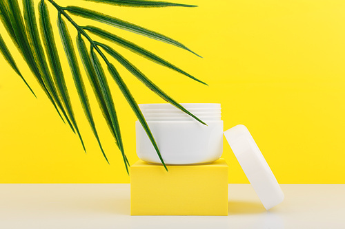 White opened cosmetic jar on yellow podium with palm leaf against bright yellow background. Plastic cosmetic jar for face mask, cream, scrub or balm