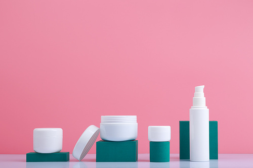 Set of cosmetic bottles on white table with green geometric podiums against pink background with space for text. Concept of skin care cosmetic set and beauty treatment