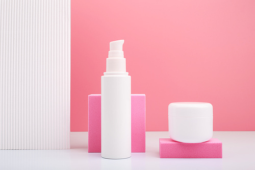 Set of white cosmetic tube and jar with face cream and under eye cream, mask or scrub against bright pink background and white wall with stripes.