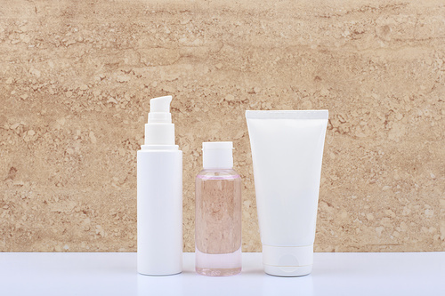 Set of beauty products for daily skin care in white unbranded tubes on white bathroom shelf against beige marble background. Concept of cosmetic products for skin cleaning, exfoliating, hydrating and anti age treatment