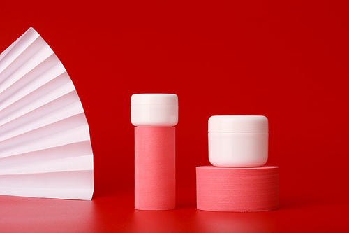 Creative set with two white cosmetic jars with skin care products on pink geometric shapes for product presentation against red background with white waver. Concept of premium or asian cosmetics