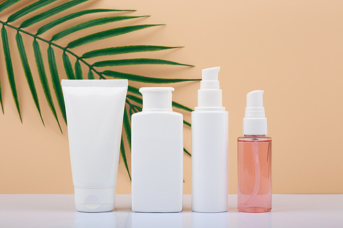 Set of white unbranded cosmetic bottles for face and body care in a row against bright beige background with palm leaf. Concept of organic, natural, eco friendly cosmetics and beauty products