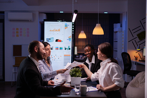 Workaholics businesspeople brainstorming financial company ideas analyzing strategy paperwork late at night in business office meeting room. Multi-ethnic coworkers checking solution