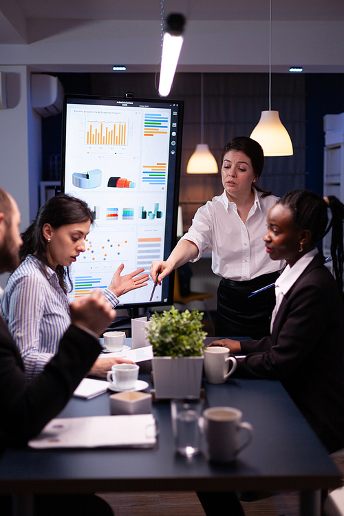 Multi-ethnic businesspeople discussing financial company solution sitting at conference table in meeting room. Diverse teamwork overworking at management strategy using tv late at night.