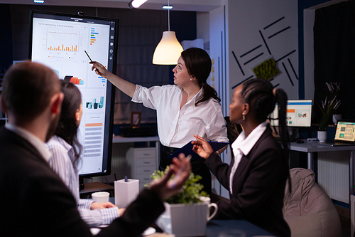 Multi ethnic businesspeople brainstorming company ideas checking graphs statistics using tv standing at conference table. Diverse teamwork working at management presentation in meeting room