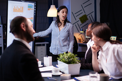 Workaholic focused businesswoman explaining management solution pointing strategy on monitor overworking in company business office meeting room. Multi-ethnic coworkers discussing ideas in evening