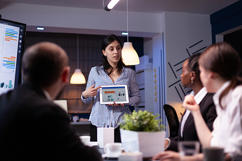 Manager holding tablet pc in meeting room late at night explaining graphs to her team. Diverse multi-ethnic teamwork solving financial strategy. Business leader holding presentation for new project