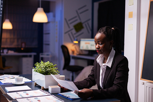 Focused workaholic african american businesswoman working at company financial charts presentation late at night in meeting office room. Overworked manager with dark skin checking profit statistics