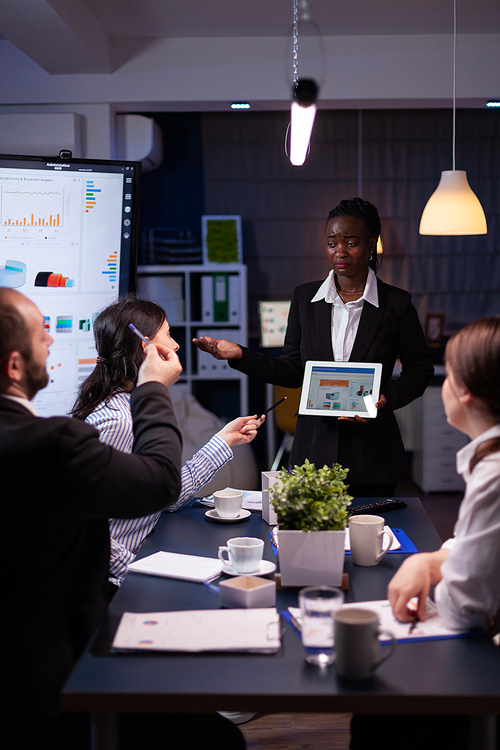 Exhausted workaholic african american showing financial graphs using tablet overworking in company office meeting room. Multi-ethnic businesspeople discussing management statistics late at night