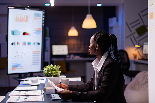 Focused workaholic african american businesswoman working at company financial charts presentation late at night in meeting office room. Overworked manager with dark skin checking profit statistics