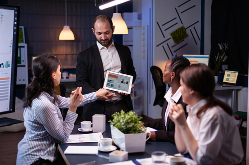 Entrepreneur businessman showing company strategy using tablet for corporate presentation working overtime in meeting office room late at night. Diverse multi-ethnic teamwork brainstorming ideas.