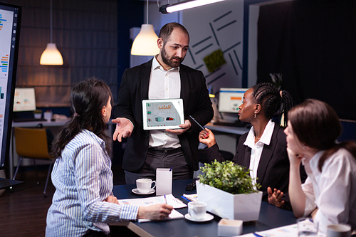 Overworked businessman showing financial graphs presentation using tablet brainstorming company ideas. Workaholic diverse multi-ethnic businesspeople overworking late at night in office meeting room