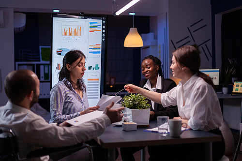 Handicapped paralyzed leder in wheelchair explaining company statistics overworking in office meeting room late at night. Diverse multi-ethnic coworkers brainstorming ideas