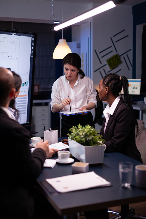 Entrepreneur woman brainstorming management strategy working hard in meeting office room late at night. Diverse multi-ethnic business team looking at financial company presentation on monitor.