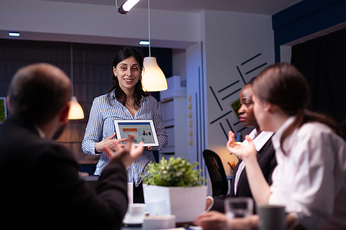 Overworked workaholic entrepreneur woman shwoing marketing graphs using tablet overworking at company solution late at night in meeting room. Diverse multi-ethnic teamwork solving financial strategy