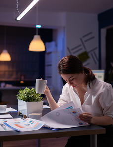 Overworked businesswoman working overtime in business company office meeting room in evening analyzing marketing charts while drinking coffee. Entrepreneur hardworking at financial profit reports