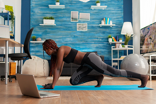 Black fit athletic woman training for muscle strenght doing mountain climbers position on yoga mat dressed in sportwear leggings, in home living room following online instructions.