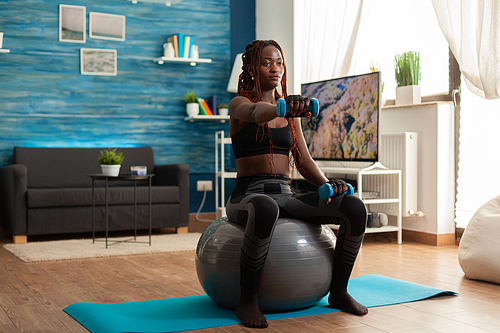 African woman using stability ball keeping outstretched arms working out shoulders using blue dumbbells, in home living room for muscle shaping and healthy lifestyle, dressed in sportwear.