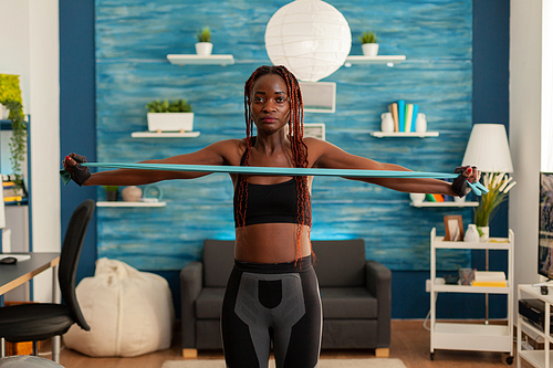 Black strong fit woman standing in living room working out shoulders using resistance band dressed in sportive sportwear. Active african exercising with rubber band.