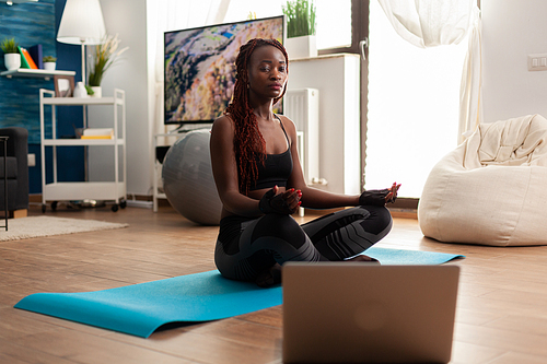 Black woman sitting on yoga mat practicing calm harmony mediting zen for healthy lifestyle, relaxing in lotus pose. Listening instructor instruction during online training.