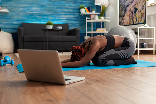 Black woman sitting on yoga mate doing stretching relaxing body after watching online training looking at laptop in home living room, on the floor for health wellbeing.