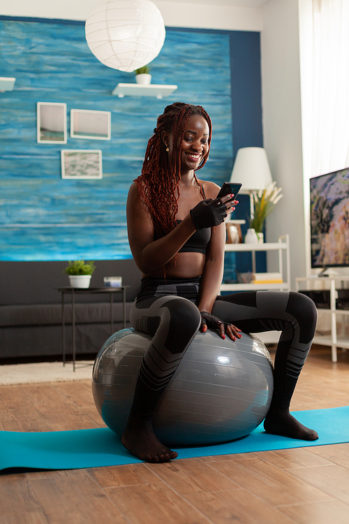 Sporty athletic black woman relaxing on stability ball checking social media using smartphone after intense workout exercising, in home living room sitting on fitness mat for muscle strenght.