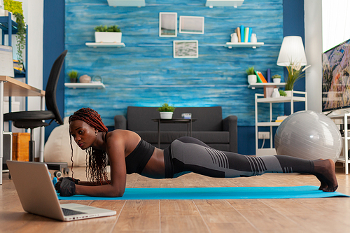 Athletic fit black woman in leggings sitting on yoga mat doing plank pose, exercising core abdominal strenght for muscle balance watching online training in home living room.