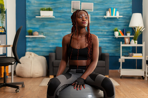 Tired fit black woman after working out in home living room, resting using stability swiss ball, wearing sportive outfit. Training indoors for muscular growth.