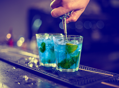 Bartender is pouring blue syrup into the glass with ice cubes on the bar counter