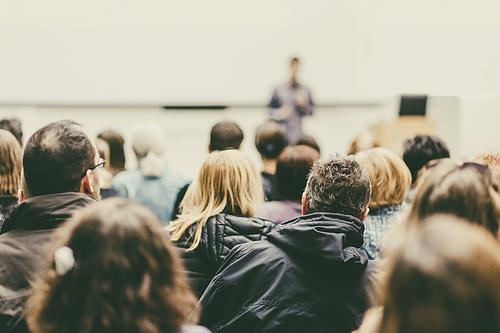 Male speaker giving presentation in lecture hall at university workshop. Audience in conference hall. Rear view of unrecognized participant in audience. Scientific conference event.