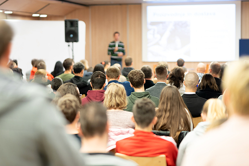 Speaker giving presentation in lecture hall at university. Participants listening to lecture and making notes.