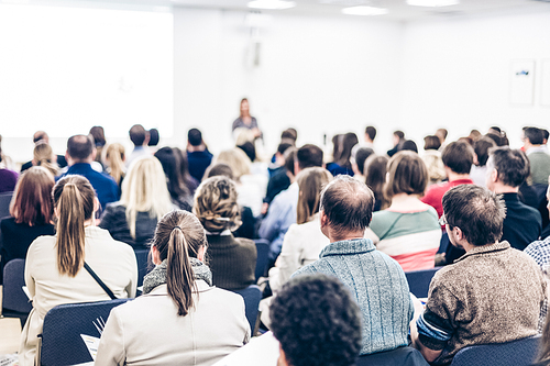 Business and entrepreneurship symposium. Female speaker giving a talk at business meeting. Audience in conference hall. Rear view of unrecognized participant in audience. Copy space on whitescreen.