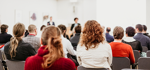 Business and entrepreneurship symposium. Female speaker giving a talk at business meeting. Audience in conference hall. Rear view of unrecognized participant in audience.