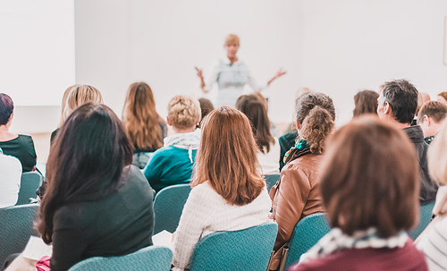 Business and entrepreneurship symposium. Female speaker giving a talk at business meeting. Audience in conference hall. Rear view of unrecognized participant in audience.