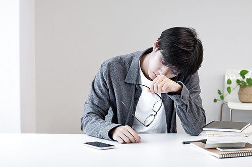책과 휴대전화를 책상 위에 둔 채 현대식 홈오피스에서 두통을 겪고 있는 젊은 아시아 대학생 남자. 편두통을 느끼는 과로한 MZ세대 한국 학생