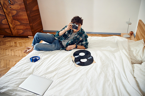 Stylish guy lying on bed with vinyl records and using retro camera stock photo