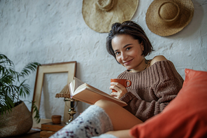 Smiling pretty lady with hot drink sitting on bed while reading