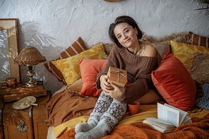 Smiling pretty lady resting with hot drink while sitting on bed