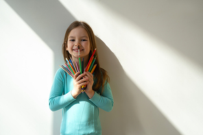 Smiling cute child with colorful drawing pencils standing against white wall stock photo. Website banner