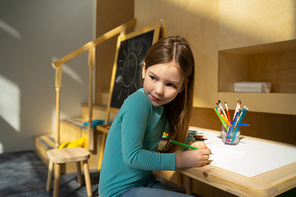 Cute little girl with pencil sitting at the table and looking over shoulder stock photo