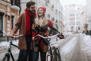 Handsome man embracing his pretty woman in winter outdoors. They are walking along the street while holding handlebars of bikes. Copy space in right side